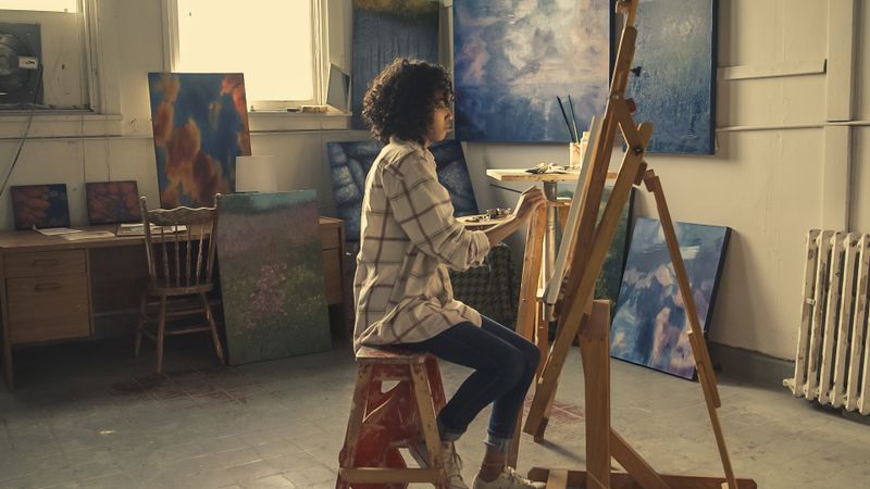 Woman with curly hair painting on a canvas easel in a sunlit art studio with abstract artworks
